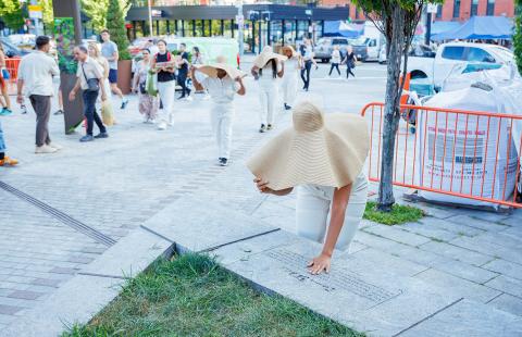 In a park, a woman in a floppy tan hat leans over and touches a dedication in stone. Behind her, more women in floppy hats walk in procession.