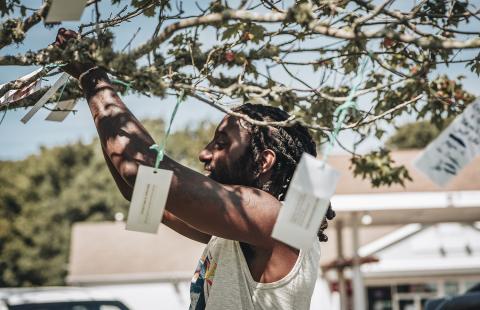 A young, Black guy ties a slip of paper to a tree with string. On the tree around him, there are other slips of paper blowing in the wind.