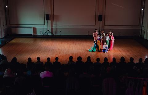 A group of performers stand in spotlights together on a half empty stage. A large crowd watches on.