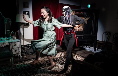 In a spotlight and on stage, a woman, in a green satin dress, dances with a person dressed as a demon.