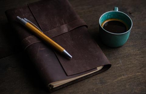 On a wooden table, a leatherbound notebook and a teal cup of coffee.