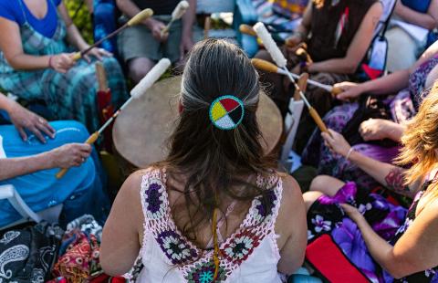 From behind, a woman, with a beaded hair tie, plays in a drum circle.