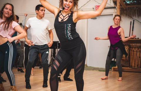 Ana, with curly hair and in an all black athletic ensemble, leads a trio of dancers while speaking into an ear piece mic.