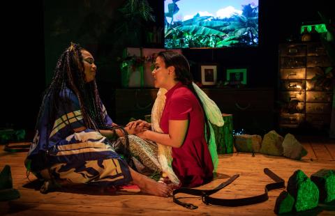 A Black woman and a Brown woman hold hands, while wrapped in blankets. They're on a stage with a projection of mountains behind them.