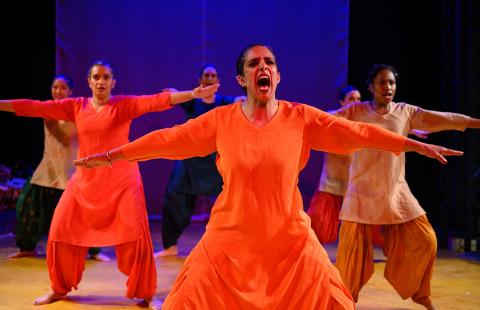 A brown woman in orange dress holds her arms out and screams. Four dancers are behind her.