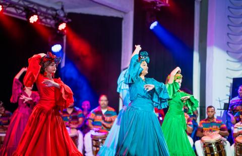 On a stage, four women in monochromatic but bright colored costumes perform. They wear headwraps that match their dresses.