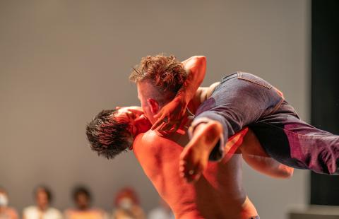 Two dancers kissing. Makisig Akin is lifted horizontally and balanced on Anya Cloud’s shoulder. Both dancers are shirtless and wearing jeans. Makisig is facing the camera with softly closed eyes. Anya is facing away from the camera.