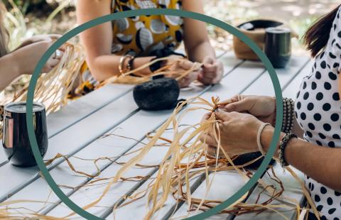 A green outline of a sphere overlaid on light-skinned women weaving hay at a picnic table.