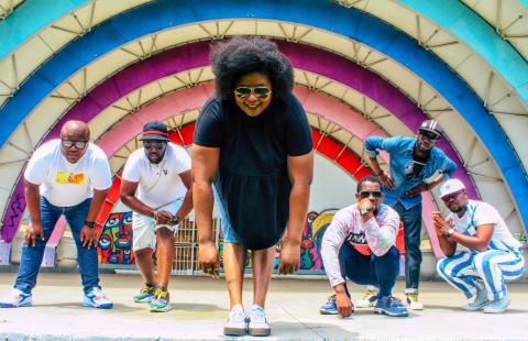 A group of 6 Black people (1 woman, 5 men) standing on a stage in what appears to be an outside amphitheater. They are crouching down towards the camera and all are wearing stylish casual clothing and cool sunglasses.