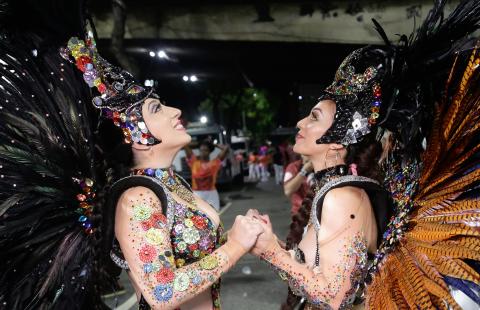 Two women hold hands and look to the ceiling. They're in feather wings and beaded carnival attire. 