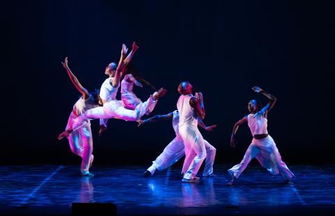On a stage, six dancers, in all white costumes, perform. Two leap in the air, one in front of the other. 