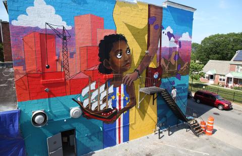 On the side of a building, a mural where a young Black girl holds a ship. A taller person, in yellow, holds her shoulder while she looks back.