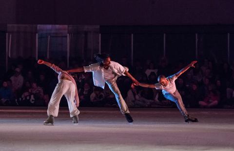 Three ice skaters slide while holding hands. One skates backwards and leans over.