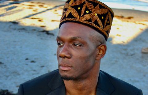 Harold is a Black man in a kufi and a black blazer. They are on the beach with the water hitting the coast behind them.