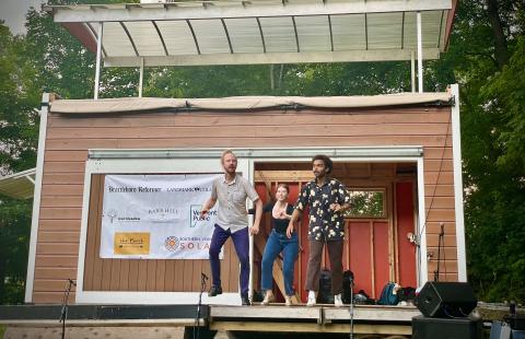 Three performers on a small outdoor stage with a wooden facade and sponsor banner.