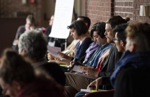 Indoors, a row of people sit and look at booklets they're holding.