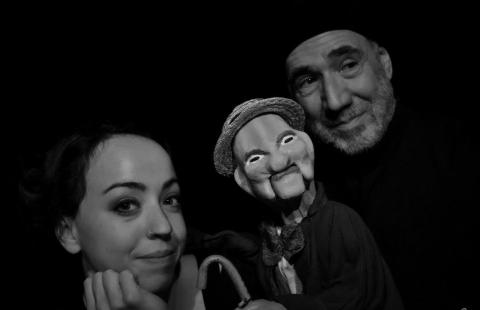 In black and white, a young white woman and an older white man, in a black beanie that fades into the background, pose with a wooden puppet.