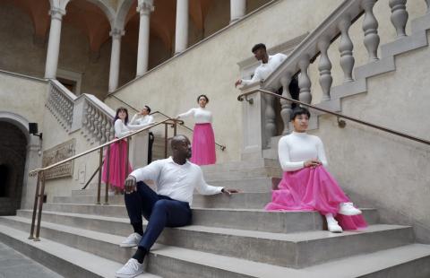 On stone steps, six dancers - men in white shirts and black slacks and women in white shirts and pink skirts - pose.
