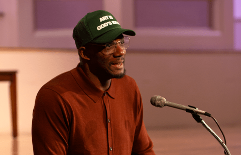 A Black man, in a trucker hat and burnt orange blouse, stands behind a podium.