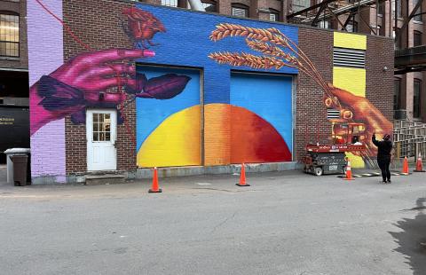 A person takes a photo, with their phone, of a mural of two hands holding grains and a flower over a sun-like orb.