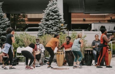Outdoors by a parking garage, a group of Black folks dance and play the drums.