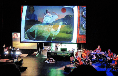 Puppeteers and an orchestra are on either side of a projection of a TV that shows a character running.