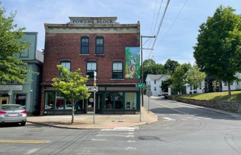 On a street corner, in a small town with no traffic or pedestrians, a mural on the side of a brick building. Below, the building has an awning. Street lines and trees frame the photo.