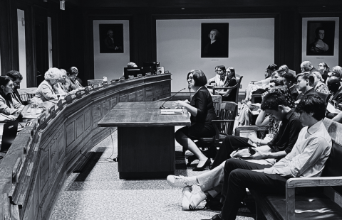 In a courtroom, a woman sits behind a table and before a group of folks.