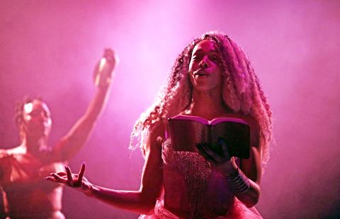 In front of a dancer and in pink light, a Black woman reads from a book.