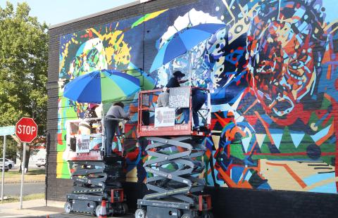 Outside, two folks, on risers, paint a mural on the side of a brick building.