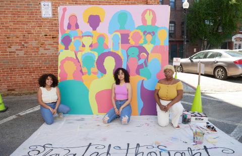 Three women of color kneel before a painting of a group of women in pastel colors.