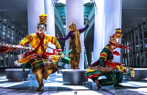 In a mall, with pillars and round, backless benches, a woman poses on a bench and two folks in ornate red, yellow, and green costumes twirl.