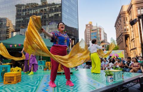 On an outdoor stage in a city, performers hold fabric and sing admist brightly colored set pieces.