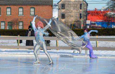 Ice skaters wear shiny silver and purple costumes and a piece of fabric they hold blows in the wind.