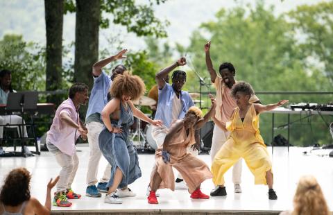 A group of 7 Black dancers on an outdoor stage; they wear soft colors and are surrounded by musicians  and leafy green trees.