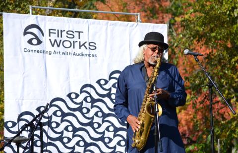 Mixashawn has long gray hair and wears a fedora. Outside and in front of a sign that says "FirstWorks," he plays the Saxophone.