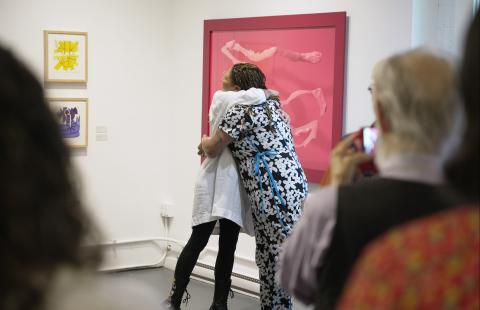 In a gallery, two women embrace with paintings on the wall behind them and a crowd clapping in the foreground.