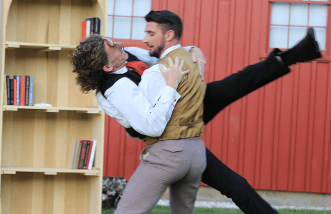 Outside, by a barn and a book shelf, two men in suits dance.