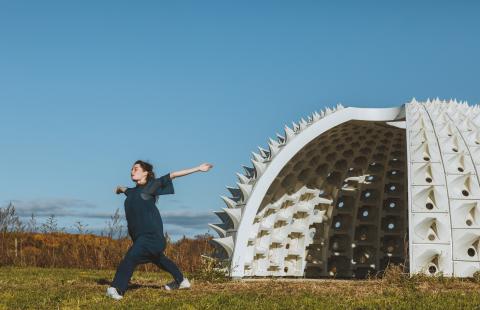 Outside, a light-skinned woman in all navy holds her arms out; she stands in front of a structure that's white and has holes and spikes in it.