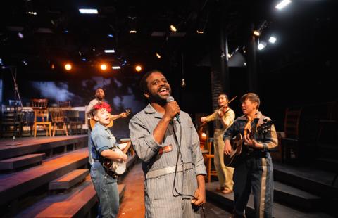 Five folks perform. A Black man sings at the center with the rest of the band behind him. There's a stage behind them to the left and steps in both directions.