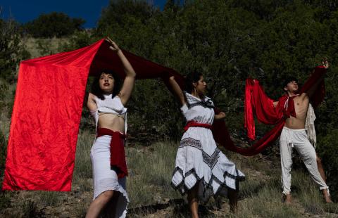 Three folks hold shiny red fabric. They wear whites and creams and pose in front of shrubs.