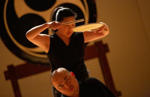 A woman holds a percussive instrument over a man's head.