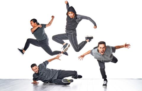 Four dancers, wearing gray, leap up, forward, or along the floor in front of a white backdrop.