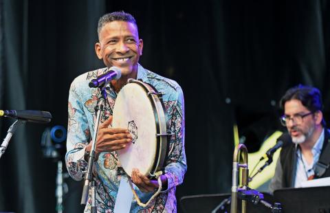 A Brown ban sings into a microphone and holds a tambourine. He wears a floral blouse.