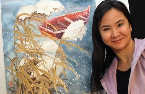 A young Chinese woman with long dark hair and wearing a lavender sweater smiles as she poses next to her painting  