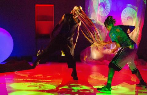 In a gallery, with projections of different colors, two dancers swap. One has a long mask on that looks like fringe.