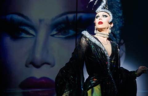 Sasha wears a sheer cloak over pants and a shimmer headpiece that snakes along her scalp. Behind her a projection of her face, which is spotlit.