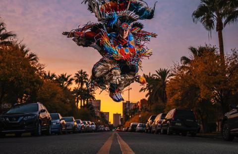 On a street, a dancer, in traditional Indigenous performance garb, jumps.