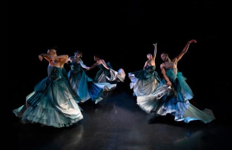 a photo features five trans, queer, and gender-expansive Sean Dorsey Dance dancers wearing elaborate floor-length blue and silver strapless ball gowns, dancing in a circle on stage. Each dancer is twirling around in a circle and has their arms out in different expressions of grace and longing. 