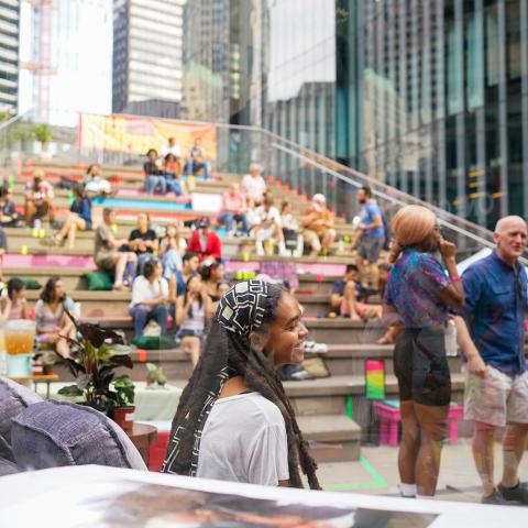 A young Black girl, with dreads wrapped in a bandana, sits outside on a blue corduroy couch. Behind her, a group of folks sit on and hang out by steps that have been decorated with chalk drawings. 